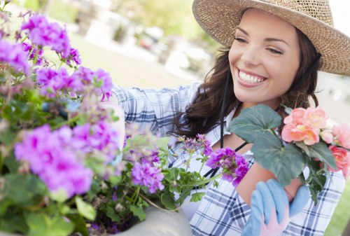 A close-up outdoor scene showing a woman with long dark hair wearing a large straw hat, smiling as she tends to flowering plants in a garden. The garden features a variety of vibrant purple, pink, and peach-colored blooms and lush green foliage. In the background, there are hints of a lawn area with well-maintained grass, a paved pathway, and trees, suggesting a neatly landscaped backyard or front garden typical of residential properties in Mortlake. The woman is dressed in a checkered shirt and gardening gloves, actively working among the flower beds, which are bordered by soil and decorative stone or brick edging. The scene appears to be set on a sunny day with natural light illuminating the plants and her face, evoking a peaceful and well-maintained outdoor space managed by professional gardening services such as Hedge Trimming Mortlake, which ensures tidy hedges and lush landscape features in the local area.