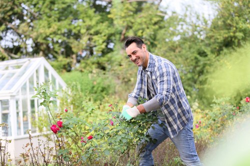 A man in casual clothing, including a blue checked shirt and jeans, is smiling while trimming a lush, green hedge with garden pruning shears in a neatly maintained outdoor garden space. The garden features a vibrant flower bed with pink and red flowers in the foreground, edged with soil and some small plants, and a well-kept lawn area with dense grass. Behind him, there are mature trees and shrubs adding natural division to the yard, with a small greenhouse structure visible on the left side. The scene is illuminated by bright, natural daylight, suggesting a clear, mild weather condition typical for outdoor gardening activities in Mortlake. This image exemplifies professional gardening and hedge trimming services that could be provided by companies like Hedge Trimming Mortlake, supporting local outdoor maintenance needs in the SW14 postcode area.