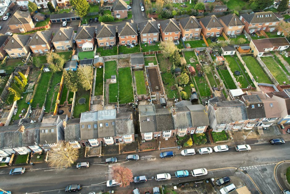 An aerial view of a residential neighbourhood showing a row of terraced houses with pitched roofs, each with small front gardens and driveways, some with parked cars. Behind the houses, there are nume